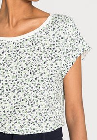 Vrouw in een kortmouwen witte top met kleine groene en zwarte bloemmotieven en een ronde halslijn, gecombineerd met donkere broek.