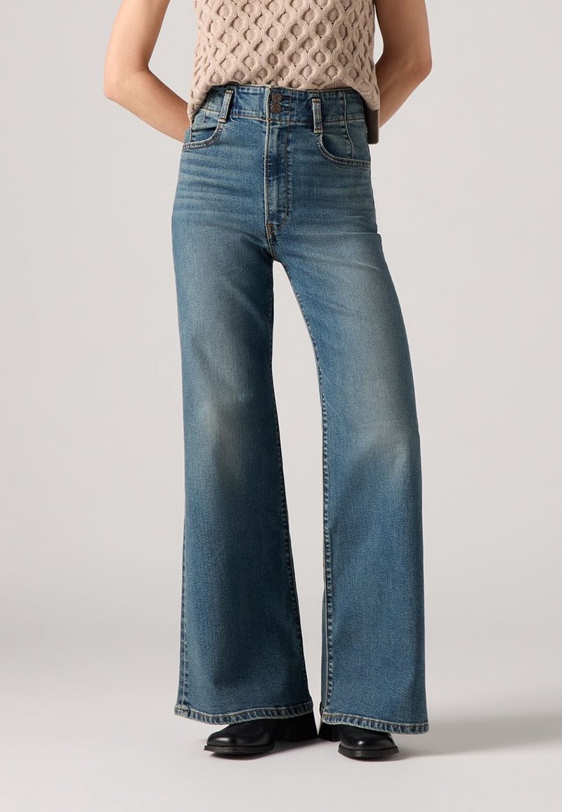 Femme portant un jean bleu taille haute à jambes larges avec un léger délavage, associé à des chaussures noires et un top beige en maille sans manches.