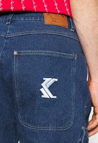 Pantalones de mezclilla con un lavado azul, que presentan una etiqueta de cuero marrón y un logo bordado en blanco en el bolsillo trasero. Incluye detalles de herrajes dorados.