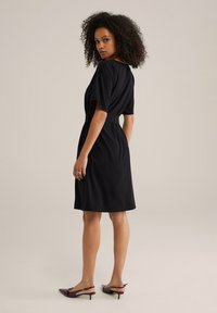 Robe noire en tissu doux, avec une coupe décontractée, des manches courtes et une taille nouée. Associée à des talons aiguilles sombres.