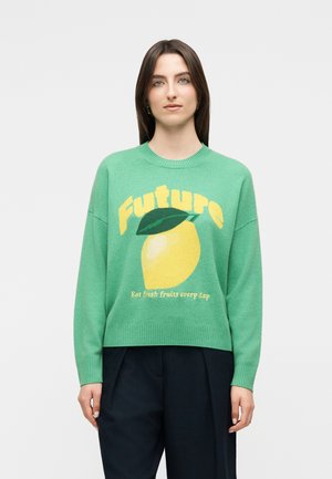 Femme aux cheveux bruns raides portant un pull vert avec un graphisme de citron jaune et le texte "Future" et "Mangez des fruits frais chaque jour."