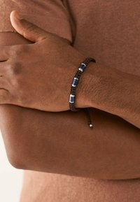 Brun vævet armbånd med tre blå rektangulære perler og sølvdetaljer. Justerbar snor med en metalperle til lukning.