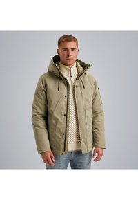 Olivovo zelená zateplená bunda s kapucňou, so zipsom a patentným zapínaním, nosená cez krémový pletený sveter. Hladká textúra, funkčný dizajn.