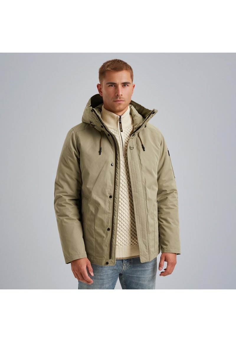 Olivovo zelená zateplená bunda s kapucňou, so zipsom a patentným zapínaním, nosená cez krémový pletený sveter. Hladká textúra, funkčný dizajn.