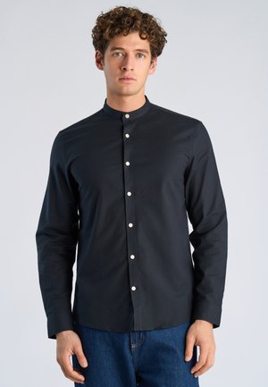 Junger Mann mit lockigem Haar trägt ein schwarzes Hemd mit Stehkragen und blaue Jeans, steht vor einem schlichten Hintergrund.