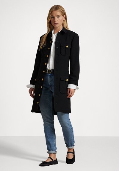 Manteau noir avec des boutons dorés, comportant quatre poches avant et une coupe ajustée. Porté sur une chemise blanche et associé à un jean bleu clair et des ballerines noires.