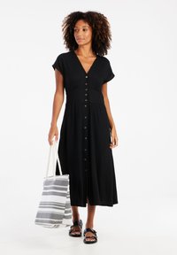 Robe midi noire à texture avec des manches courtes et des boutons sur le devant. Le mannequin tient un sac fourre-tout rayé et porte des sandales noires.