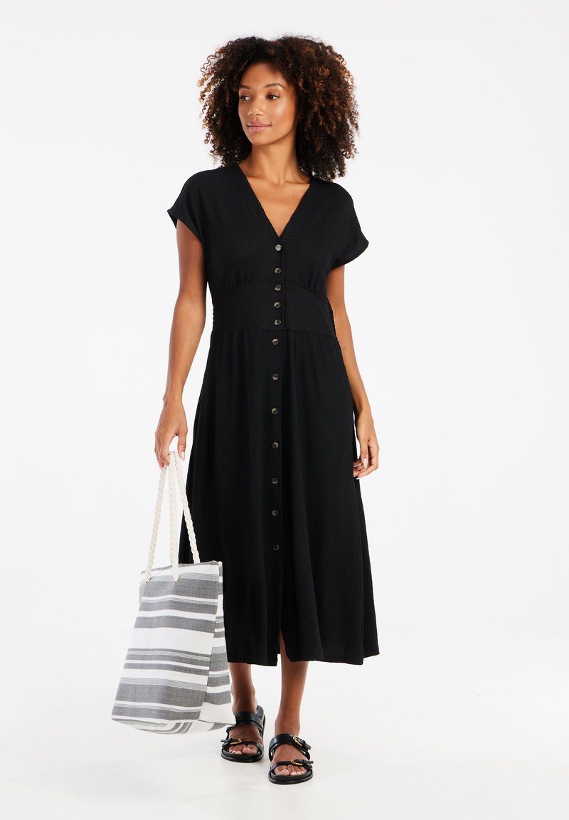 Robe midi noire à texture avec des manches courtes et des boutons sur le devant. Le mannequin tient un sac fourre-tout rayé et porte des sandales noires.
