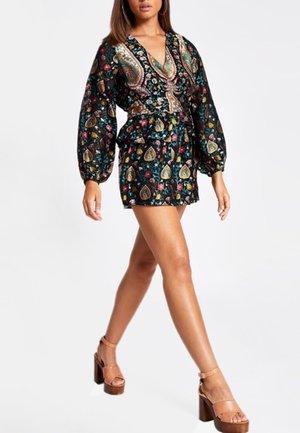 Vrouw die loopt, gekleed in een korte jurk met lange mouwen, bedrukt met bloemen en paisley, gecombineerd met bruine plateauhakken.