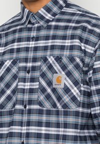 Gekleurde geruite overhemden in marineblauw, wit en oranje; met een kraag, twee borstzakken en een Carhartt-logo patch. De textuur lijkt glad.