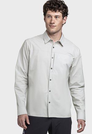 Mann trägt hellgraues langärmeliges Hemd mit kleinem Logo auf der Brust und dunkle Hose, steht vor einfachem Hintergrund.