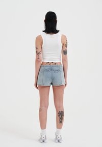 Top corto blanco y pantalones cortos de mezclilla azul claro con un logo bordado en la parte posterior; zapatos planos; tatuajes visibles en las piernas.
