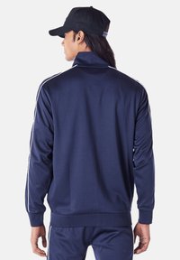 Marineblaue Trainingsjacke mit hohem Kragen, langen Ärmeln und weißen Seitenstreifen. Verfügt über eine glatte Textur und enganliegende Bündchen, getragen mit einer schwarzen Mütze.