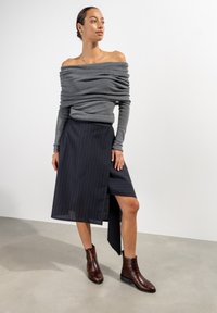 Grauer Off-Shoulder-Pullover, marineblau gestreifter Wickelrock und braune Stiefeletten mit strukturierter Oberfläche. Schlichte, elegante Formen und Linien.