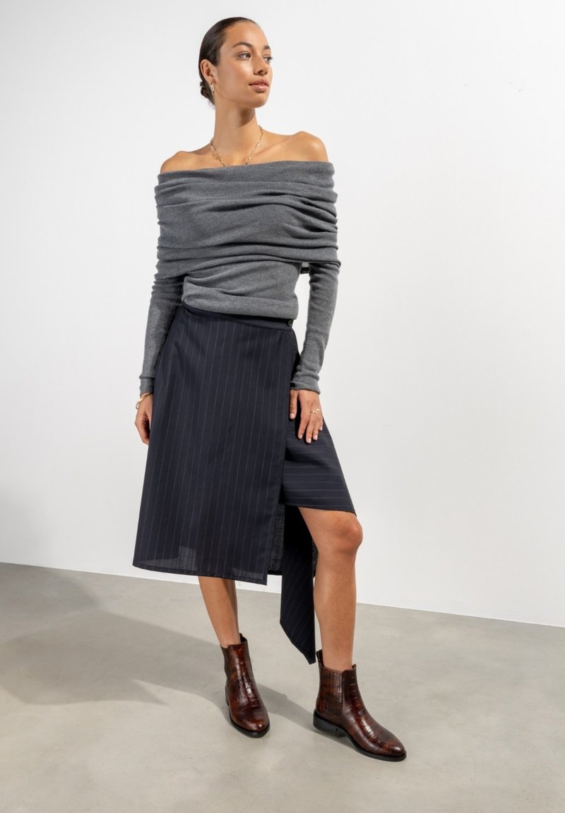 Grauer Off-Shoulder-Pullover, marineblau gestreifter Wickelrock und braune Stiefeletten mit strukturierter Oberfläche. Schlichte, elegante Formen und Linien.