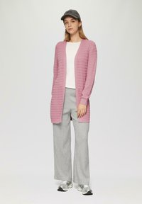 Lange roze gebreide cardigan met een zigzagpatroon, open voorkant, gedragen over een witte top en grijze wijde broeken met patroon, gecombineerd met sneakers.