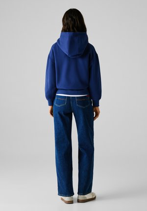 Felpa blu con cappuccio e orlo a costine, abbinata a jeans blu a vestibilità ampia. Una camicia bianca sbuca sotto la felpa. Calzature casual.
