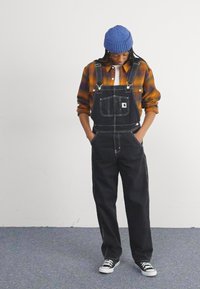 Svarte denim overaller med lomme foran, kombinert med en brun og oransje rutete skjorte, og en blå ribbestrikket lue. Svarte joggesko fullfører stilen.