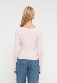 Γυναίκα με μακριά ξανθά μαλλιά, φορώντας ένα ανοιχτό ροζ μπλουζάκι με μακριά μανίκια και μπλε τζιν, γυρισμένη με την πλάτη σε λευκό, απλό φόντο.