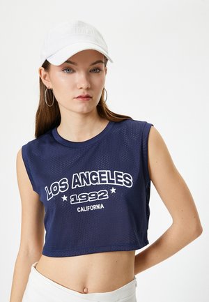 Marineblå cropped tank top med mesh stof, med hvid trykt tekst "LOS ANGELES 1992 CALIFORNIA" og uden ærmer. Hvid kasket bæres.