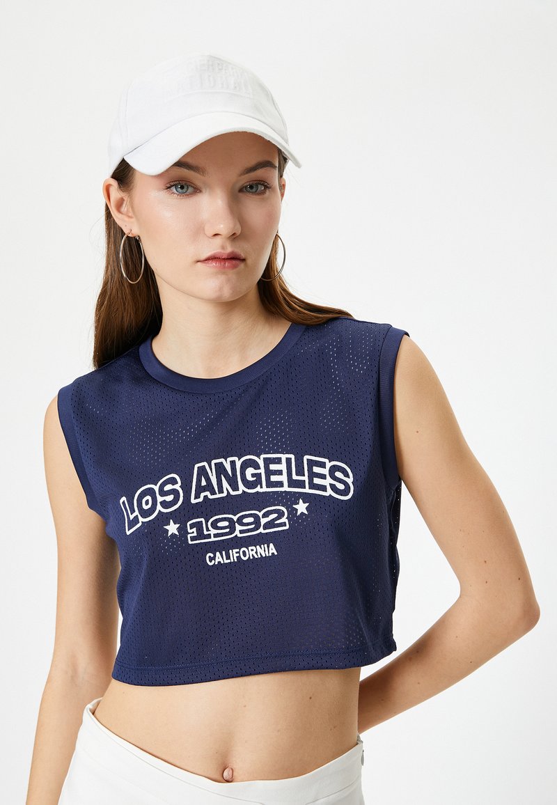 Granatowy krótki top na ramiączkach z siateczkowej tkaniny, z białym nadrukiem "LOS ANGELES 1992 CALIFORNIA" i bez rękawów. Na głowie biała czapka.