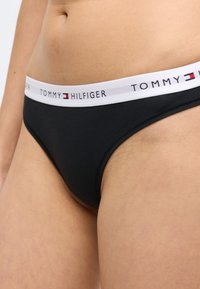 Chilot tanga negru din bumbac, prevăzut cu o bandă elastică albă pe talie, marcată cu "TOMMY HILFIGER" în negru și roșu. Textură netedă, design clasic.