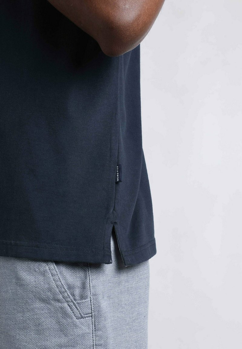 Navy blue cotton T-shirt with side vents and a small label. Worn with light gray textured shorts. Close-up of hem and side seam.