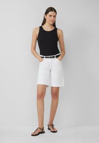 Schwarzes geripptes Tanktop, weiße knielange Shorts mit Umschlägen, schwarzer Gürtel mit Metallschnalle und schwarze flache Sandalen, vor einem grauen Hintergrund präsentiert.