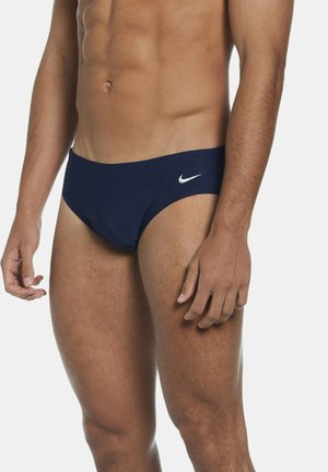 Primo piano della parte inferiore del torso di un uomo che indossa slip da nuoto Nike blu scuro su uno sfondo chiaro uniforme.