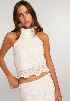 Joven mujer con cabello largo y castaño, vistiendo un crop top sin mangas de encaje blanco con estampado floral y una falda a juego sobre un fondo sencillo.