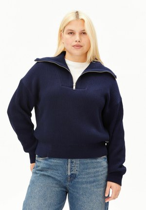 Femme blonde portant un pull bleu marine à demi-fermeture éclair sur un haut blanc et un jean bleu, debout avec une main sur la hanche.