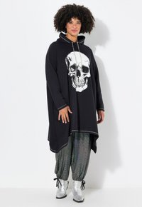 Zwarte hoodie jurk met een grote witte schedelafbeelding, voorzien van een capuchon, lange mouwen en zilveren hardware, gecombineerd met iriserende broek en zilveren laarzen.