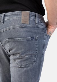 Graue Denim-Jeans mit einer glatten Textur, ausgestattet mit einem Lederetikett am Bund und einem kleinen blauen Etikett an der Gesäßtasche.