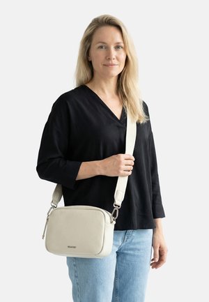 Donna con capelli biondi che indossa una maglietta nera e jeans blu tiene una borsa a tracolla in pelle beige con una tracolla bianca regolabile.
