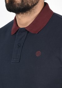 Námornícky modrá polo tričko s vínovou golierom, obsahujúce dvojitý gombík a malé červené logo zemegule na ľavej hrudi. Textúrovaná tkanina.