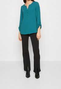 Blusa teal a maniche lunghe con scollatura a V e tre bottoni. Abbinata a pantaloni neri svasati con spacchi, completati da stivali neri alla caviglia.
