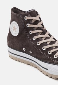 Brązowe zamszowe sneakersy typu high-top z beżowymi sznurowadłami, białą gumową podeszwą i czarnymi akcentami; na boku znajduje się logo w kształcie gwiazdy.