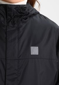 Giacca nera realizzata in materiale leggero e resistente all'acqua. Presenta un cappuccio, un tessuto testurizzato e una piccola etichetta con logo sul petto.