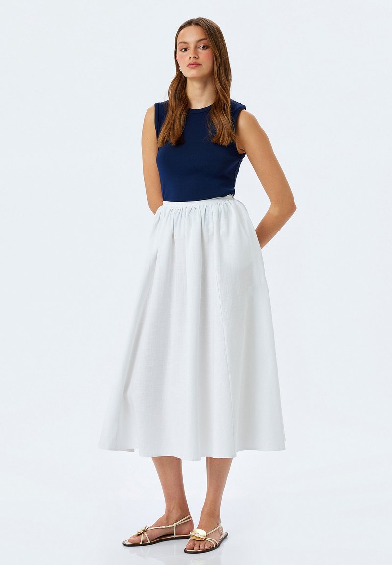 Donkerblauwe tanktop gecombineerd met een witte, knielange, geplooide rok. De outfit heeft een aansluitend lijfje en vloeiende stof. Gouden sandalen.