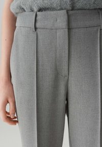 Primo piano di una persona che indossa pantaloni grigi a vita alta con passante per cintura e frontale plissettato, insieme a un top grigio con texture.