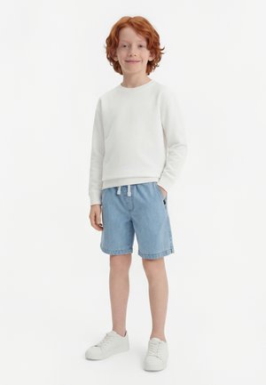 Rødhåret barn står, iført en hvid langærmet skjorte, lyseblå denimshorts, hvide sneakers, smilende med den ene hånd i lommen.