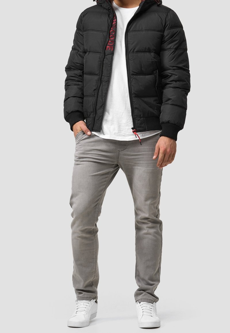 Homme portant une veste matelassée noire, un t-shirt blanc, un jean gris clair et des baskets blanches, debout avec une main dans la poche sur un fond gris.