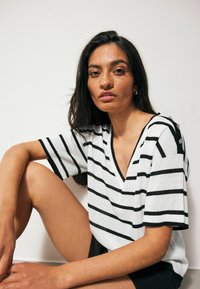 Striped V-neck sweater in white and black, with short sleeves and button accents on the shoulder, worn with black shorts. Smooth texture.