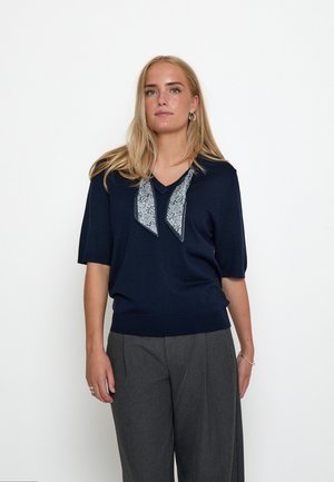 Donna con lunghi capelli biondi che indossa una maglia blu navy a maniche corte con dettaglio di sciarpa fantasia e pantaloni grigi a gamba larga su sfondo bianco.