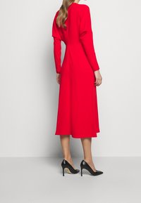 Vestido rojo de manga larga hecho de un tejido suave, con un corpiño ajustado y una falda midi acampanada, combinado con tacones de aguja negros.