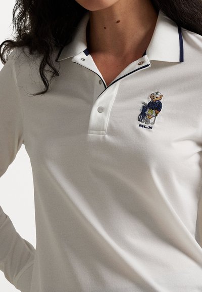 Witte polo met marineblauwe bies, voorzien van een beren borduring, twee knopen bij de kraag en een gestructureerde stof.