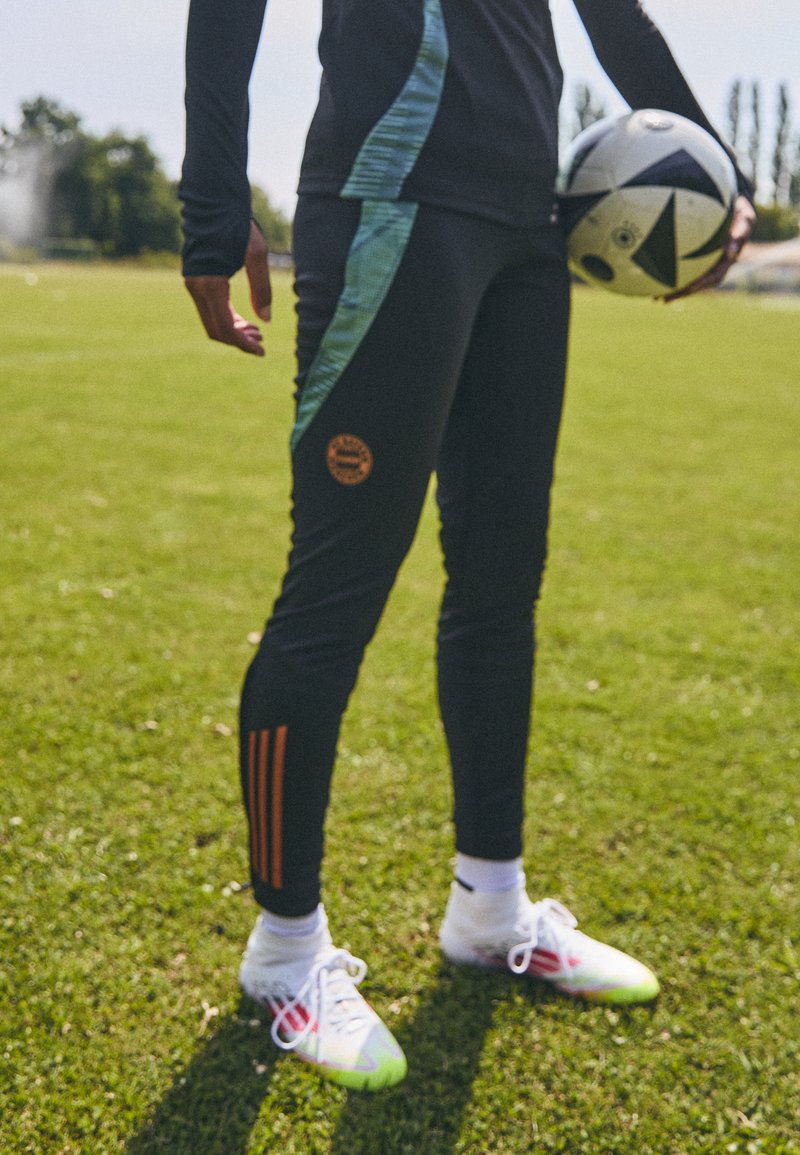 Čierne športové nohavice s teal bočnými pruhmi a oranžovým logom, doplnené bielymi futbalovými kopačkami so zelenými akcentmi, držiac čierno-biely futbalový balón.