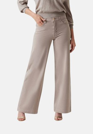 Donna che indossa pantaloni beige a vita alta e gamba larga e tacchi aperti beige, con una mano in tasca e un maglione beige.