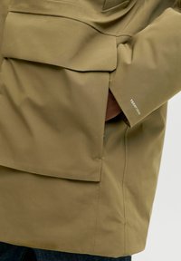 Alyvuogių žalios spalvos vandeniui atsparus švarkas su dideliu priekinio kišeniu, lygiu paviršiumi ir "TECHFUSION" logotipu ant rankovės.