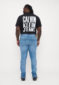 Camiseta negra con gran texto en blanco "CALVIN KLEIN JEANS" en la parte posterior, combinada con jeans skinny de color azul claro y zapatillas negras.